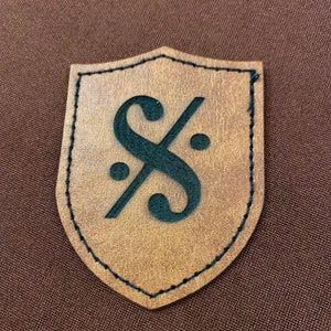 May include: A brown leather patch in the shape of a shield with a black musical note symbol inside.