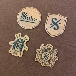 May include: Four brown leather patches with black printed designs. The patches feature a musical note, a beetle, a shield with a musical note, and a musical note with a decorative frame.