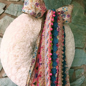 May include: A white, fluffy wreath decorated with a large bow and colourful ribbons. The bow has floral embroidery and sequins. The ribbons include pink pom-poms and turquoise floral lace, creating a festive and decorative piece.