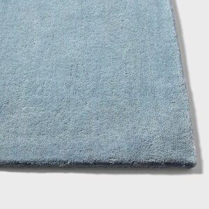 May include: A close-up of a section of a light blue rug. The rug has a soft, plush texture and a solid color. The edges of the rug are a darker shade of blue. The rug is a home decor item.