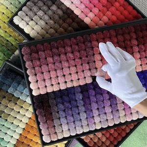 May include: A collection of colorful pom-pom samples arranged in black trays. The samples display a gradient of colors, ranging from greens and yellows to reds and purples. A gloved hand is touching the samples.