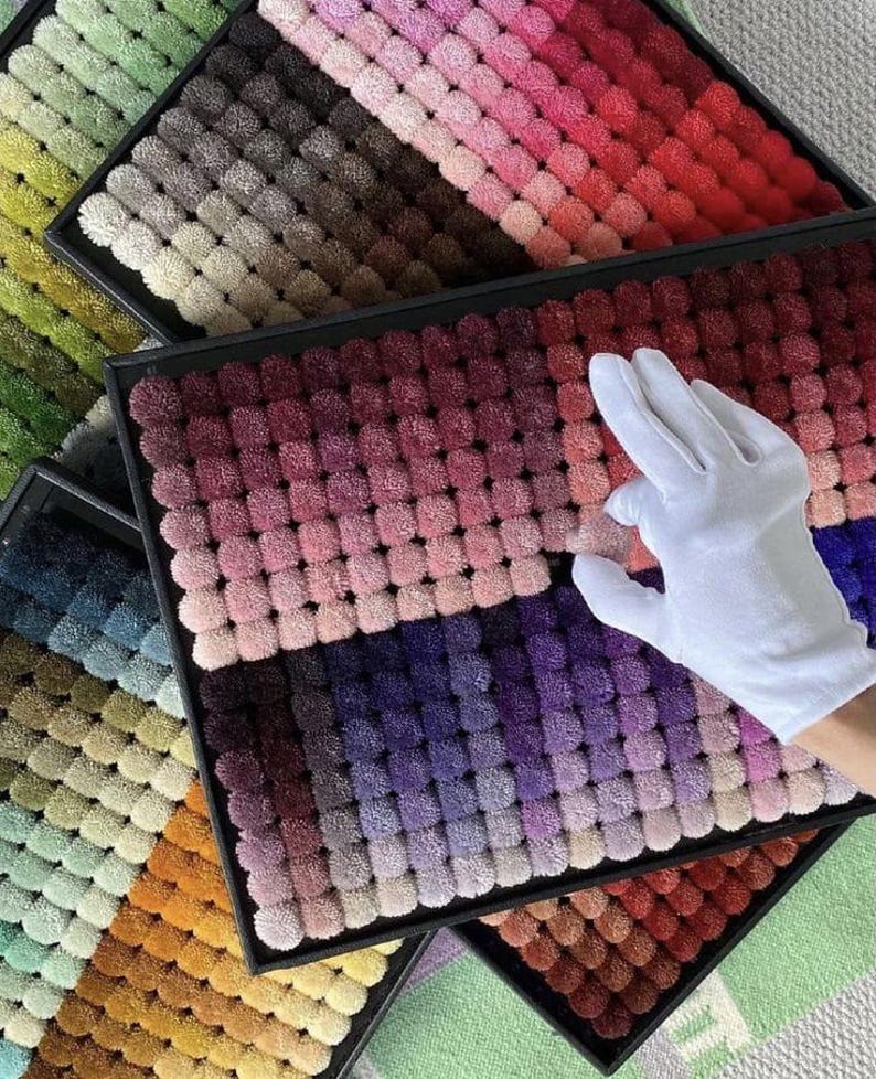 May include: A collection of colorful pom-pom samples arranged in black trays. The samples display a gradient of colors, ranging from greens and yellows to reds and purples. A gloved hand is touching the samples.