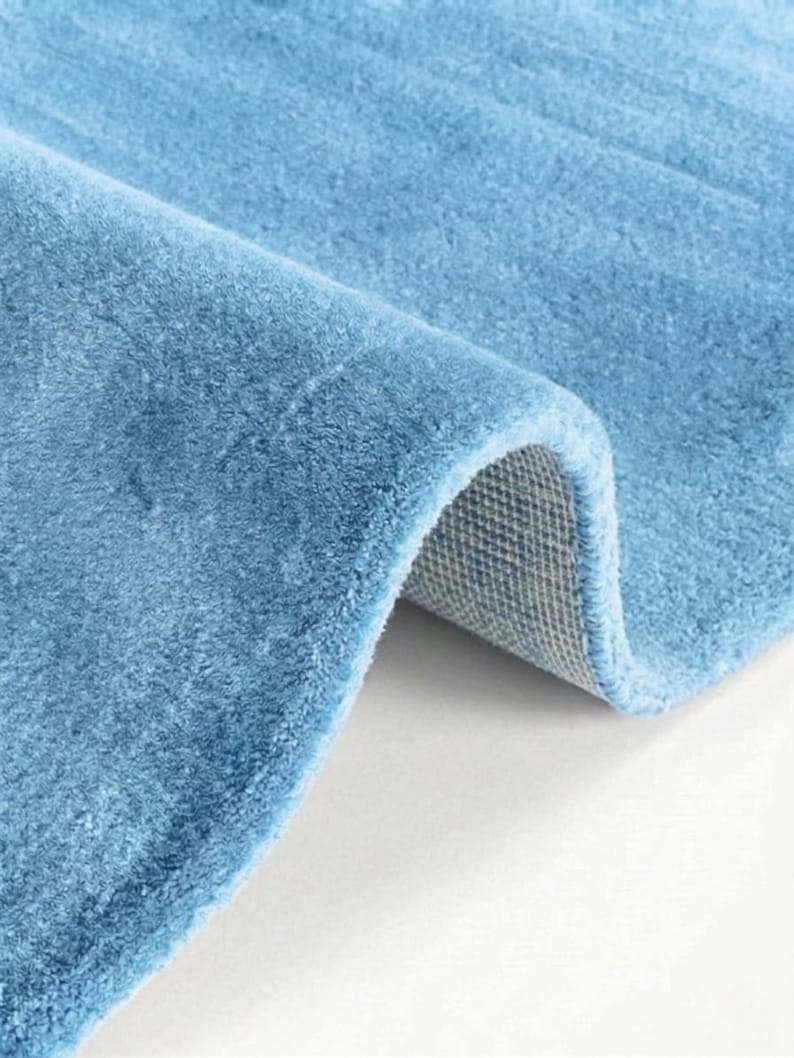May include: A close-up of a rolled-up, light blue rug. The rug has a soft, plush texture and a visible backing. The color is a solid, muted blue, and the rug appears to be made of a thick, durable material. The rug is likely for home decor.