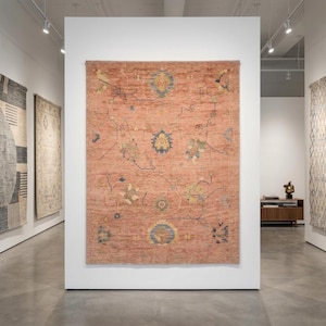 May include: A large, rectangular, peach-coloured rug with an intricate pattern of blue, yellow, and green floral and geometric designs. The rug is displayed on a white wall in a gallery setting, with other rugs visible in the background.