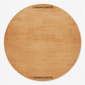 May include: A large, circular rug in a light brown shade, featuring a wood grain pattern. The text "LUKMANLOOMTEX" is printed in black at the top and bottom. The rug is set against a white backdrop.