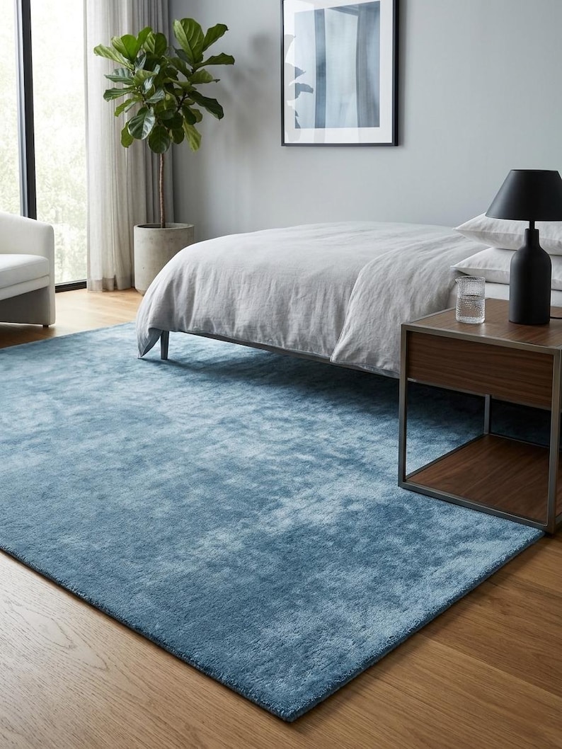 May include: A rectangular, light blue rug on a hardwood floor in a bedroom setting. The rug is the focal point, with a soft, textured appearance. A bed with light gray bedding, a side table, and a potted plant are also visible.