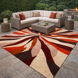 Puede incluir: Escena de patio al aire libre con una alfombra grande y colorida con un diseño de rayos de sol en tonos de rojo, naranja, marrón y beige. Un sofá seccional con cojines neutros y cojines decorativos se encuentra en un patio de piedra, con una mesa de centro y una sombrilla.