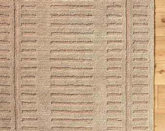 Puede incluir: Alfombra rectangular beige con una superficie texturizada y un patrón geométrico de líneas horizontales y verticales. La alfombra está hecha de un material suave y tiene una paleta de colores neutros, adecuada para varios estilos de interior.