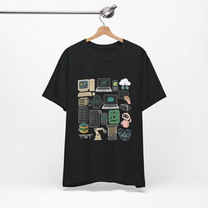 Puede incluir: Camiseta negra con una colorida ilustración de elementos tecnológicos y de IA. El diseño incluye ordenadores, portátiles, un robot, un gráfico de cerebro y otros iconos relacionados con la tecnología. La camiseta está colgada de una percha de madera.