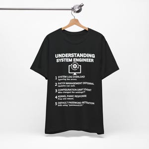Könnte beinhalten: Schwarzes T-Shirt mit weißem Text: "UNDERSTANDING SYSTEM ENGINEER" über einer Computergrafik. Darunter nummerierte Punkte, die Probleme von Systemingenieuren detaillieren, darunter "SYSTEM LOG OVERLOAD". Das T-Shirt hängt an einem Holzbügel.