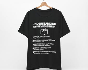 Könnte beinhalten: Schwarzes T-Shirt mit weißem Text: "UNDERSTANDING SYSTEM ENGINEER" über einer Computergrafik. Darunter nummerierte Punkte, die Probleme von Systemingenieuren detaillieren, darunter "SYSTEM LOG OVERLOAD". Das T-Shirt hängt an einem Holzbügel.