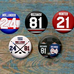Puede incluir: Cinco botones redondos con temática de hockey con nombres y números de jugadores. Los botones presentan los nombres "HOLLANDER", "ROZANOV" y "HUNTER", junto con los números "24", "81" y "21". Los colores son azul, rojo, blanco y negro.
