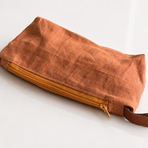 Könnte beinhalten: Ein rechteckiges Etui aus strukturiertem, rostfarbenem Stoff mit einem senfgelben Reißverschluss an der Oberkante. Eine kleine Schlaufe aus dunkelbraunem Leder ist an der Seite des Etuis befestigt und dient als praktischer Tragegriff.