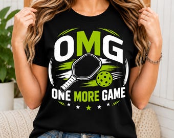 May include: A black short-sleeved t-shirt featuring a graphic design with the text OMG ONE MORE GAME in white and lime green. The design includes a black pickleball paddle and a lime green pickleball with holes, accented by white stars.