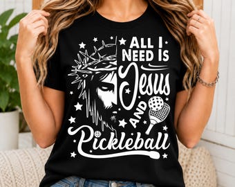 May include: A black t-shirt featuring a white graphic print. The design shows a split image of a face wearing a crown of thorns on the left and the text "ALL I NEED IS JESUS AND PICKLEBALL" on the right, accompanied by a pickleball paddle and ball.