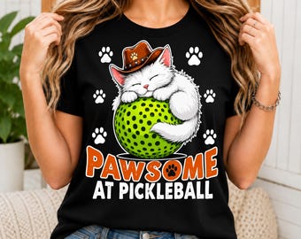 Può includere: Una t-shirt nera con la grafica di un gatto bianco che indossa un cappello da cowboy marrone e dorme su una pallina da pickleball verde brillante. Il design include impronte di zampe bianche e il testo arancione e bianco PAWSOME AT PICKLEBALL.