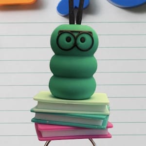 Puede incluir: Un llavero verde con forma de oruga y gafas negras se encuentra sobre una pila de libros en miniatura de colores pastel. El llavero tiene un anillo plateado y un dije decorativo.