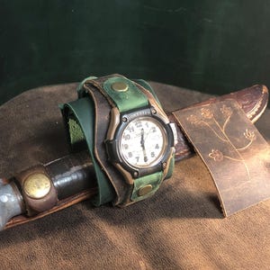 May include: A handcrafted leather watch band with a green and brown color scheme, featuring a watch with a white face and black numerals. The watch is paired with a knife and a decorative leather piece with an embossed floral design.