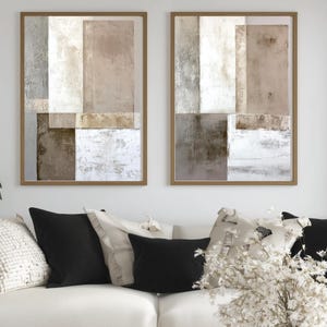 May include: Two framed abstract art prints. Each print features a geometric design with rectangular shapes in shades of beige, grey, and white. The frames are a light brown colour. The prints are displayed above a white sofa with black and cream-coloured cushions.