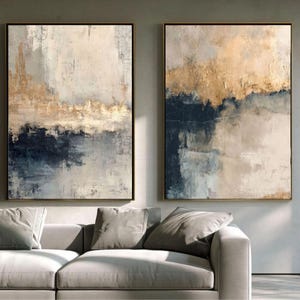 May include: Two rectangular wall art pieces featuring abstract layers of navy blue, cream, and metallic gold paint. The textured brushstrokes create a horizontal composition with a dark base, a lighter middle section, and a shimmering gold top.