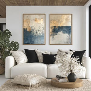 May include: Two framed abstract paintings with gold, white, and navy blue tones hang above a white sofa with black and cream pillows. A round wooden coffee table holds a black vase with white flowers.
