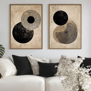 May include: Two framed abstract art prints. Each print features a beige background with overlapping black circles and concentric rings. The frames are a warm brown color. The artwork is displayed above a white sofa with black and cream pillows.
