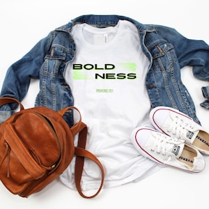 Pode incluir: Camiseta branca com as palavras "BOLDNESS" em preto e verde com o texto "PROVÉRBIOS 28:1" por baixo. A camiseta está colocada sobre uma superfície branca com uma jaqueta de ganga, uma mochila de couro castanho e ténis brancos.