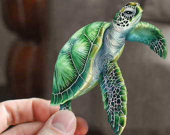 以下が含まれることがあります： 小さな緑色のウミガメで、茶色の甲羅と黒い目があります。カメは手に持たれており、背景はぼやけていて、ソファと木製のテーブルが見えます。