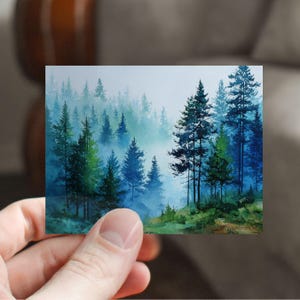 May include: A watercolor painting of a forest scene. The artwork depicts tall evergreen trees in shades of green and blue, with a misty, atmospheric background. The painting is held in a hand.
