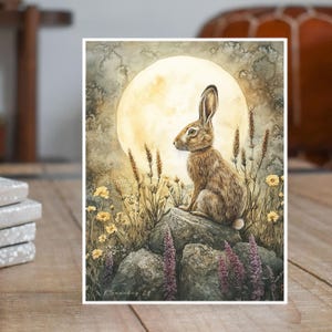 May include: A watercolor painting of a brown rabbit sitting on a rock under a full moon. The rabbit is looking to the left, and the moon is behind it. There are wildflowers and grasses in the foreground.