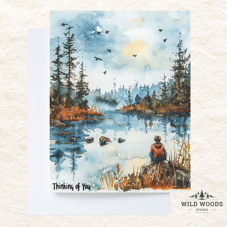 May include: A watercolour painting of a tranquil lake scene with a solitary figure seated on a rock by the water's edge. The sky is a soft blue with a few white clouds and the trees are a dark green. The water is a light blue with reflections of the sky and trees. The text "Thinking of You" is written in black at the bottom of the image.