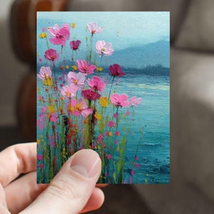 May include: A small, rectangular painting featuring pink and yellow flowers in front of a teal body of water. The flowers have varying shades of pink and are set against a backdrop of a blue mountain range. The artwork is held by a person.