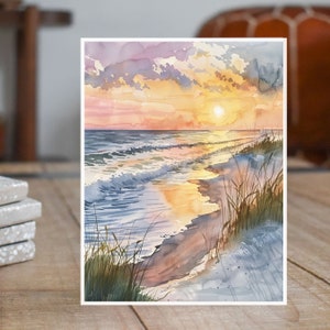 May include: Watercolor painting of a sunset over the ocean with waves crashing on the shore. The sky is a mix of pink, orange, and yellow, and the water is a deep blue. The beach is sandy and there are some green grasses in the foreground.