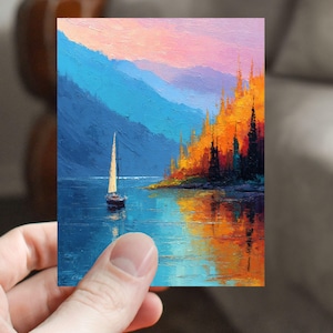 May include: A small, rectangular painting depicts a sailboat on a lake, with mountains in the background. The water reflects the sky and the trees on the right. The sky transitions from pink to blue. The painting is held by a hand.