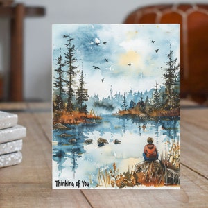 May include: Watercolor painting of a forest scene with a lake and a lone figure sitting on a rock. The sky is blue and cloudy, and the trees are green and brown. The text "Thinking of You" is written at the bottom of the image.