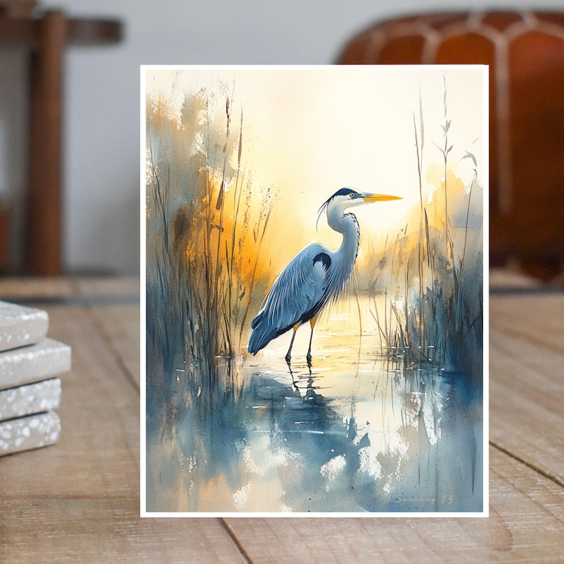 May include: A watercolor painting of a blue heron standing in a marsh with tall reeds and a reflection in the water. The heron is facing to the right and has a long, thin beak. The background is a soft, muted yellow and blue.