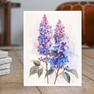 May include: Watercolor painting of lilac flowers in shades of purple, blue, and pink. The flowers are arranged in a bouquet with green leaves and stems. The background is a soft watercolor wash of pink and blue.
