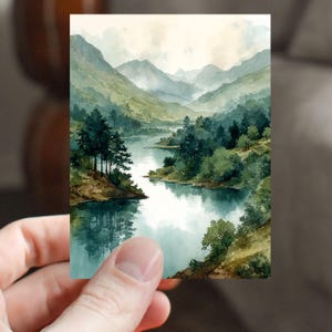 May include: Watercolor painting of a serene mountain lake scene. The artwork depicts a calm lake surrounded by lush green trees and rolling hills. The sky is overcast, creating a soft, diffused light over the landscape. The painting is held by a person.