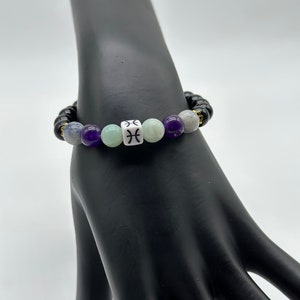 May include: A black, purple, and green beaded bracelet with a white bead featuring the Pisces zodiac symbol.