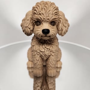 Puede incluir: Figura de caniche de color canela con un pelaje rizado y texturizado. El perro tiene ojos oscuros y una nariz negra. La figura está de pie, mostrando sus detalles y diseño. El fondo es de un color suave y neutro.