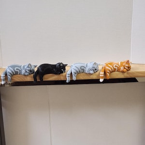 May include: Four cat figurines in various colors: gray with white stripes, black, gray with white stripes, and orange with white stripes. The cats are lying down, with their bodies draped over the edge of a wooden surface. The background is a plain white.