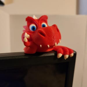 Pode incluir: Figura de dragão vermelho impressa em 3D, com olhos azuis e dentes brancos. O dragão está empoleirado em cima de um monitor de computador preto, adicionando um toque lúdico. Objeto decorativo.