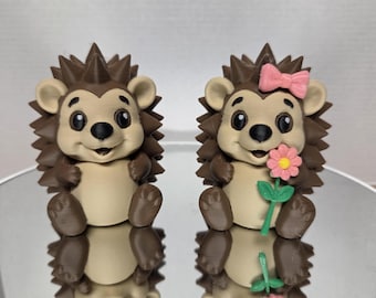 May include: Two small hedgehog figurines with brown spines and tan faces. One hedgehog has a plain head, while the other features a pink bow on its head and holds a pink flower with a green stem. Both figures have black eyes and noses.