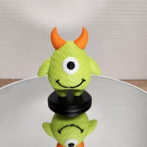 May include: A small, plush, lime green monster toy with one eye, orange horns, and a smiling mouth. The toy has small arms and is standing on a black base. The toy's reflection is visible below.