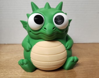May include: A green, cartoon-like dragon figurine with large white eyes and a tan belly. The dragon has a friendly expression and is sitting down. The figurine is made of a solid material and has a smooth finish.