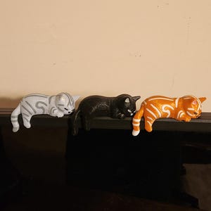 Op de afbeelding: Drie decoratieve kattenbeeldjes op een rij. Eén is wit met grijze strepen, de middelste is zwart en de laatste is oranje met witte strepen. De katten liggen, met hun poten en staarten over de rand.
