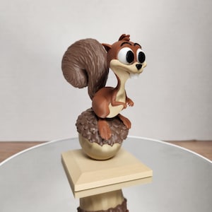 May include: A brown cartoon squirrel figurine with a large, fluffy tail, perched atop a detailed acorn. The acorn sits on a light brown square base. The squirrel has large eyes and a toothy grin.