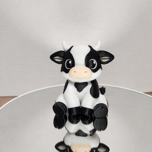 Puede incluir: Figura artesanal de una vaca sentada, con un diseño de manchas blancas y negras. La vaca tiene un hocico de color canela claro, orejas negras y pequeños cuernos. La figura se refleja en una superficie plateada brillante.