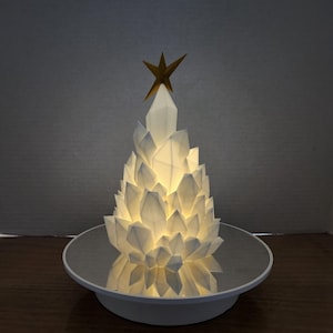 以下が含まれることがあります： 白い幾何学的なクリスマスツリー型のランプで、上部に金の星が付いています。 ランプは内側から照らされ、温かい光を放ちます。 光と木の形を反射する鏡面の上にあります。 ベースは白です。