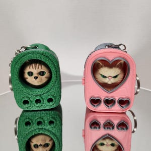 May include: Two cat-themed keychains. One is green, the other is pink. Each keychain features a cat's face peeking out of a carrier. The pink carrier has a heart-shaped window and heart cutouts. Both have silver key rings.